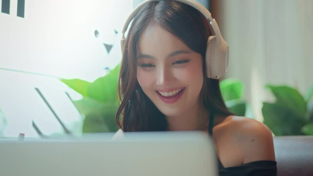 Cheerful asian woman wearing wireless headphones sitting at cafe table, studying online, watching webinar and taking notes on laptop &ndash; digital nomad lifestyle and distant education