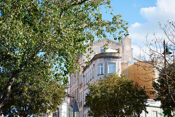 A view of a cityscape featuring buildings partially obscured by lush green trees. The sky is bright with scattered clouds, creating a vibrant atmosphere.