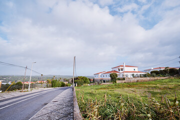 Obraz premium A rural road with a sidewalk, bordered by a grassy field and houses under a cloudy sky.