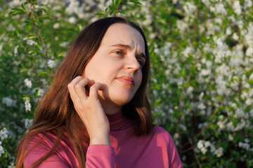 Young woman scratches her itchy skin near flowering trees. Reacting to pollen or skin irritation...