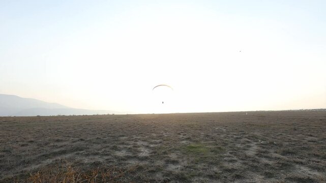 Lone powered paraglider gracefully flies across a hazy, atmospheric sky during sunset. Below, distant silhouetted hills create a serene, minimalist backdrop for this adventure sport.