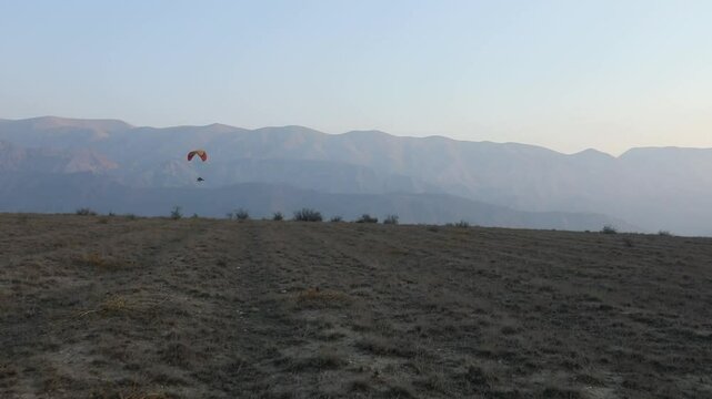 Lone powered paraglider gracefully flies across a hazy, atmospheric sky during sunset. Below, distant silhouetted hills create a serene, minimalist backdrop for this adventure sport.