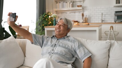 Obraz premium Happy grandfather and grandmother making video call with their son at home. Elder man holding smart phone and sitting at sofa while old woman greeting with cheerful vibe at modern house. Myrmidon.