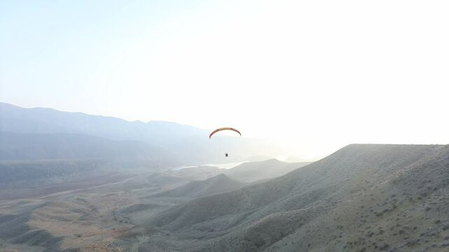 Lone powered paraglider gracefully flies across a hazy, atmospheric sky during sunset. Below, distant silhouetted hills create a serene, minimalist backdrop for this adventure sport.