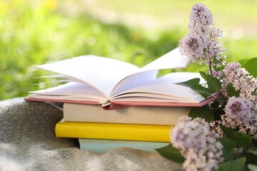 Stacked books, lilac flowers and blanket on green grass outdoors, closeup