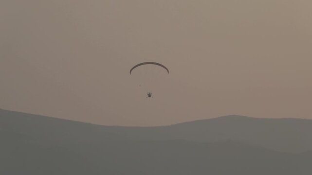 Lone powered paraglider gracefully flies across a hazy, atmospheric sky during sunset. Below, distant silhouetted hills create a serene, minimalist backdrop for this adventure sport.