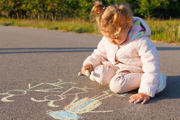 Young girl kneels drawing figures with chalk on pavement, showing childhood creativity, outdoor fun, and temporary street art.