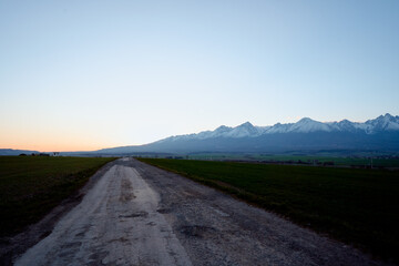 Serene Rural Road Leading Towards Majestic Snow-Capped Mountain Range at Sunrise..