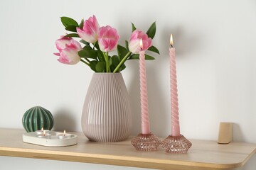 Elegant holders with burning candles, tulips in vase and other decor on wooden table near white wall