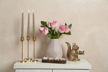 Elegant candlesticks with burning candles, tulips in vase and other decor on dresser near beige wall