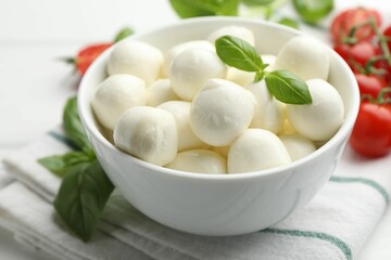 Tasty mozzarella cheese balls in bowl, basil and tomatoes on table, closeup