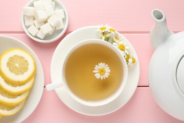 Delicious chamomile tea in cup, fresh flowers, sugar cubes and lemon on pink wooden table, flat lay