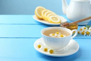 Delicious chamomile tea in cup, fresh flowers, lemon and honey on light blue wooden table, closeup. Space for text