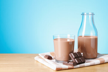 Tasty chocolate milk in glass, bottle and pieces on wooden table against light blue background, space for text