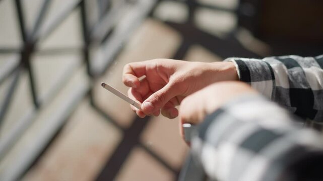 hand view of smoker tapping lit cigarette against iron railing twice to drop ash in warm sunlight with blurred urban background checkered shirt sleeve flickering in breeze