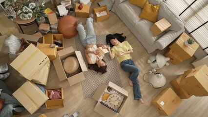 Top down aerial view of couple lesbian lying down at living room floor surrounded with cardboard box while sharing story in book. Happy lover spending time together and laughing together. Myrmidon.