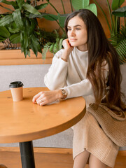 Elegant woman with coffee sitting in cafe, surrounded by green plants, wearing neutral toned stylish outfit
