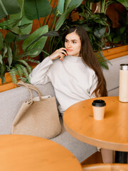 Elegant woman with coffee sitting in cafe, surrounded by green plants, wearing neutral toned stylish outfit