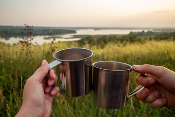 Two hands holding stainless steel camping mugs in a toast against a scenic sunset landscape with rolling hills and a winding river symbolizing outdoor adventure travel and shared moments in nature.