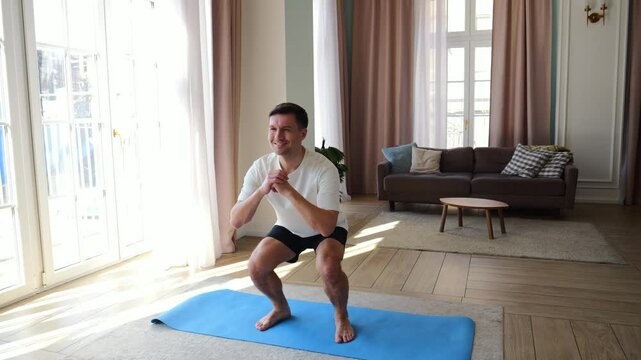 A man is exercising with squats on a soft mat in a bright, sunny indoor space.