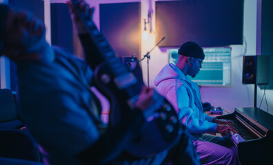 Creative musicians work together in a recording studio. One plays guitar while another focuses on a keyboard. The atmosphere is vibrant and innovative, highlighted by colored lighting and equipment.
