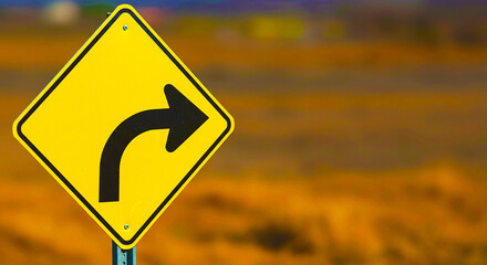 A yellow road sign with a right turn arrow against a blurred background of brown and orange hues