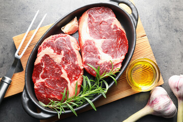 Pieces of raw beef meat with spices in baking dish, oil and fork on gray textured table, flat lay