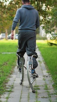 Back shot of guy walking with stilts wearing gray hoodie and pants on paved road surrounded by green grass and trees, showcasing balance, agility, and motion in peaceful outdoor setting
