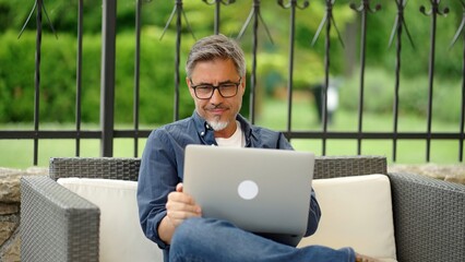 Man sitting on outdoor sofa in garden working with laptop computer in home office, managing...