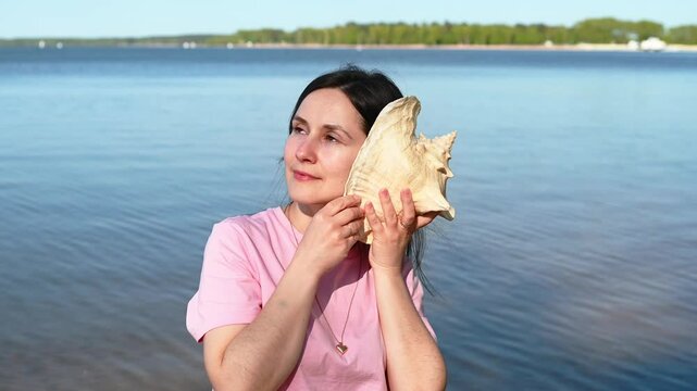 Young woman with loose hair on a lake shore holding a large seashell to her ear, dreaming of the sea, peaceful and contemplative moment in nature