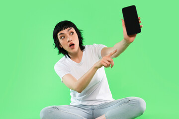 Young woman sitting on green background points at smartphone with surprise and excitement while...