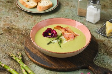 Fresh asparagus puree soup with asparagus pieces and fried bacon in a pink ceramic plate on a green concrete background. Recipes asparagus.