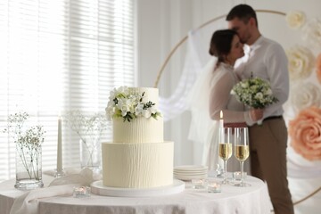 Happy married couple together indoors, focus on wedding cake and glasses of champagne on table
