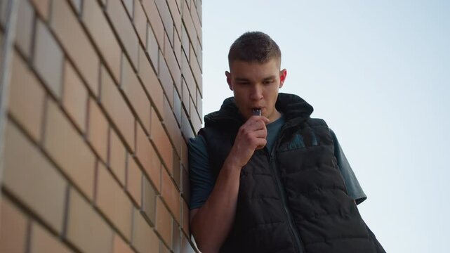 young boy leaning against brick wall outdoors inhaling shisha smoke into mouth as he coughs looking at vape device wearing black puffer vest over dark shirt in sunlight casting shadows on bricks
