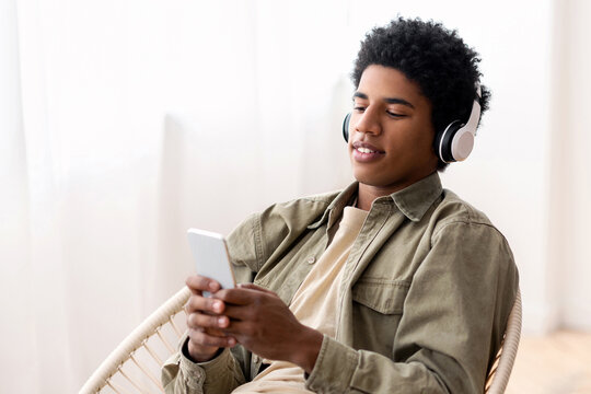 Handsome black teen in headphones choosing musical playlist on smartphone at home. Attractive African American guy enjoying great song, listening to audiobook or radio, indoors