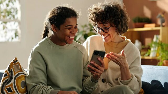 AI animated shot of happy curly-haired Caucasian mom spending time with teenage adopted African American daughter, both smiling while looking at smartphone screen, sharing precious moments