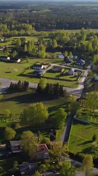 Vertical drone pull away reveals Katlakalns spring residential area with green fields, country roads, traffic, and houses surrounded by trees under clear sky and golden afternoon light