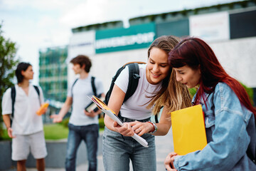 Two female university students smiling and helping each other with projects outdoors on campus