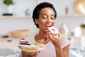 Young black woman on diet having cheat meal day, holding plate of sweets, stuffing her mouth with...