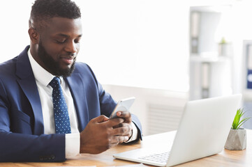 Obraz premium Handsome african businessman using cellphone at workplace, sending message or email, empty space