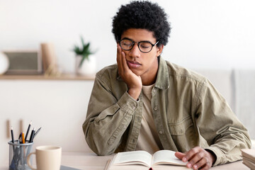 Remote education concept. Tired black male student sleeping at table while reading textbook...