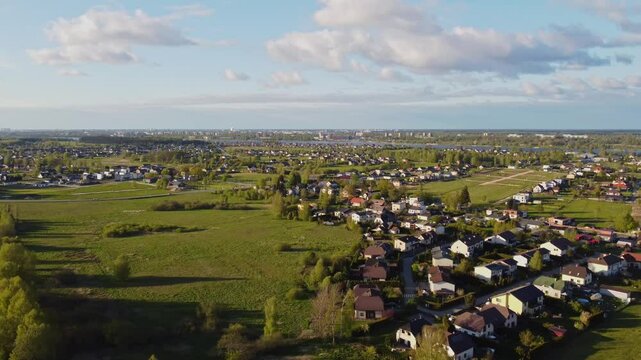 Drone slowly pulls away above Katlakalns residential area showing houses, spring green fields, tree lines, and Riga skyline on the horizon under scattered clouds and afternoon light