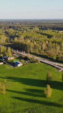 Vertical drone shot captures Katlakalns countryside with green fields, scattered houses, traffic moving on road, and dense forest in background under warm sunlight, aerial footage