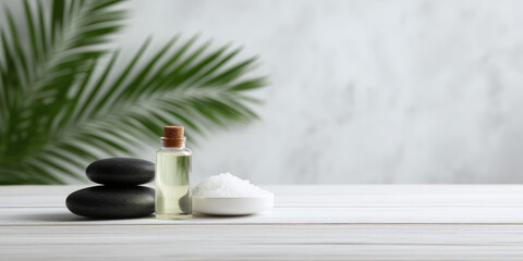 Smooth black stones, a small bottle of oil, and a dish of salt are arranged on a light wooden surface with a palm leaf in the background. Spa relaxation concept