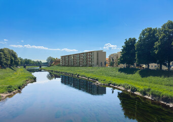 Scenic view of the Murg river in Rastatt, Germany with modern residential buildings on the right bank and lush greenery reflecting in the calm water under a bright blue sky