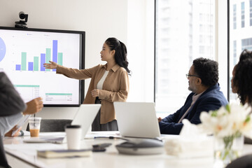 Diverse team members listen young Asian woman professional gives presentation, pointing on monitor displaying diagrams, engaged in data analysis during briefing event in boardroom. Business analytics