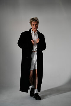 Handsome young man in black coat and shorts posing on grey background