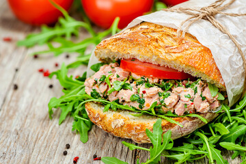 Close up of a tuna salad baguette sandwich with arugula and tomato wrapped in paper on wood table