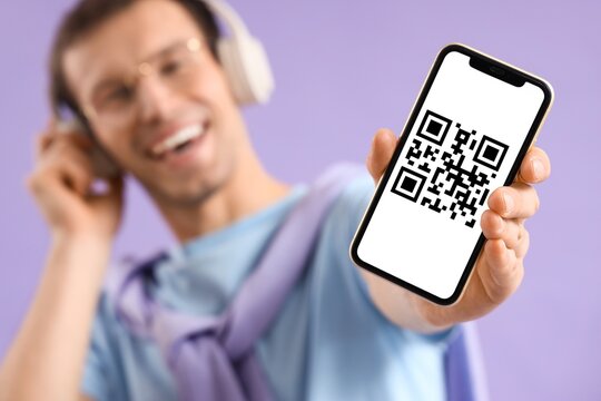 Young man holding smartphone with QR code on screen and listening to music against lilac background, closeup