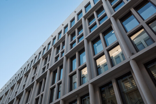An upward view of a contemporary office building facade featuring large reflective windows framed by light concrete structures and vertical columns. 